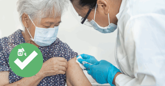 Photo of a doctor wearing a mask, lab coat, and rubber gloves administering a flu vaccine to an elderly patient wearing a mask. There is a green and white icon of a vaccination bottle and syringe and check mark in the bottom corner.