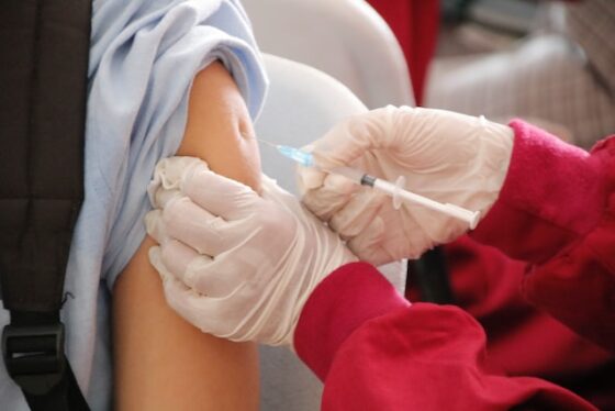 A student is being injected with a vaccine by a gloved physician with red sleeves. 