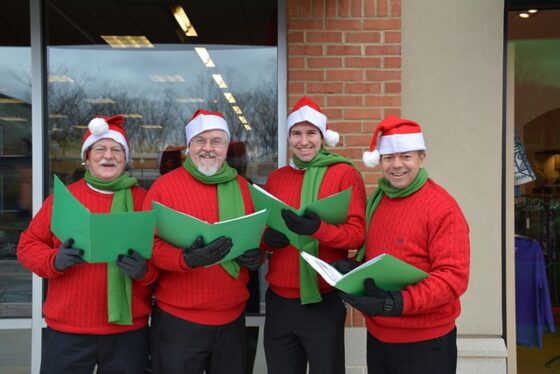 Photo of four people dressed in matching red sweaters, green scarves, red and white caps, and black mittens holding green folders full of carols. They are standing in front of a store front with large smiles on their faces. 