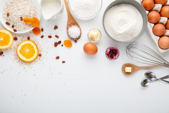 Photo of a white counter top with carton of eggs, bowl of flour, bowl of sugar, pitcher of milk, variety of spoons, some containing salt and butter. Orange and apricot slices nearby. It looks like a space to start cooking/baking. 