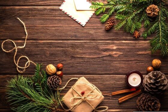 Photo of wood table with scattered cinnamon sticks. pine cones, evergreen branches, nuts, twine, and a brown paper package tied up with string. 
