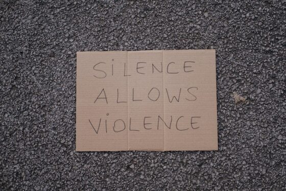Photo of a cardboard sign on pavement. This phrase is handwritten in pen on the cardboard "Silence Allows Violence" it is a call to not stay silent but instead to report. 