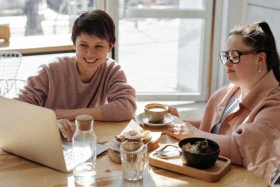 Two professional women having a meeting at a cafe. The table is covered with delicious looking food, pitchers of water, coffee, and the computer laptop with their current project. One women has short dark hair and is wearing a pale rose colored sweater. The other women has her long hair in an updo, wearing glasses and a blush blazor; she has down syndrome. 
