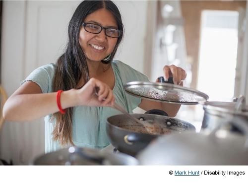Photo of a woman with dark long hair and glasses smiling while cooking and stirring the contents of a pot while she lifts the lid with the other hand. She is a woman with disabilities. Photo purchased from Disabilityimages.com