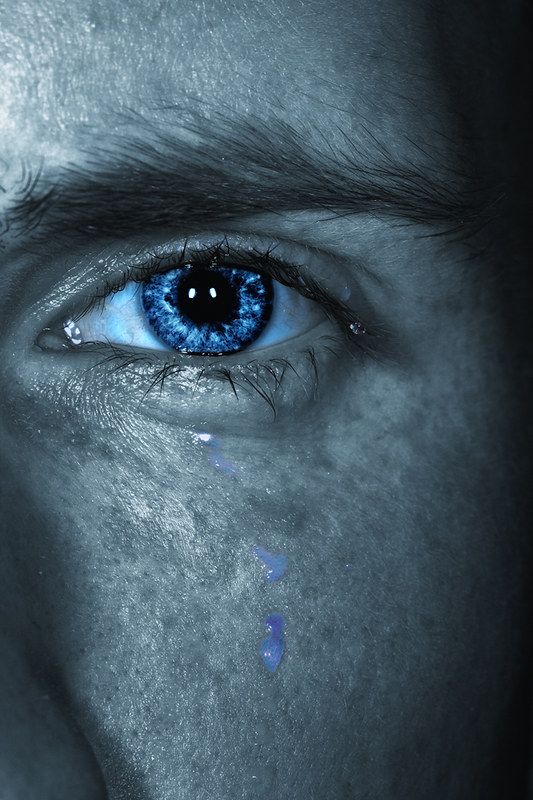 Photo closeup of a person's eye with blue tears falling. The image has been artistically altered so that the face and eyebrows are in grayscale but the eye's iris is radiant blue. 