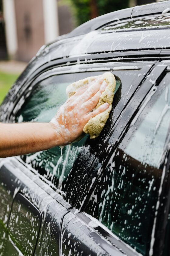 Photo of a man's Caucasian hand washing a dark colored vehicle with a yellow sponge and lots of soapy water