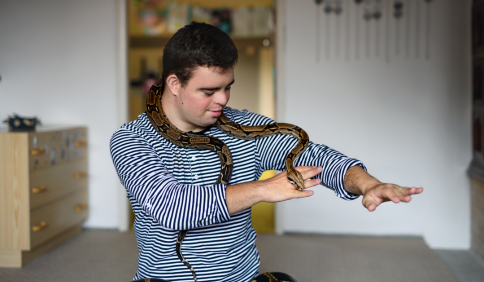 Photo of a young man with an intellectual disability wearing a long sleeve striped shirt and holding a pet snake over his shoulders and arm.