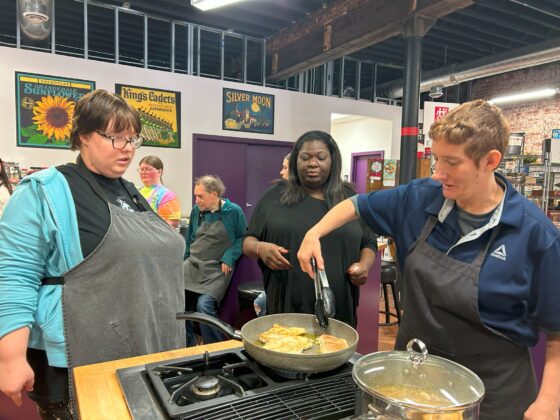 Attendees at Thyme in Your Kitchen, everyone is wearing aprons. One flips chicken in the skillet with tongs while the others oversee. 