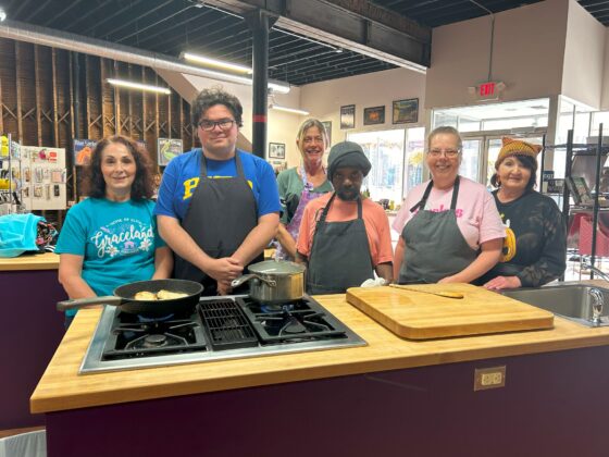 Group at a station in Thyme in Your Kitchen pose and smile for the camera together while the soup and chicken cook on the stove. 