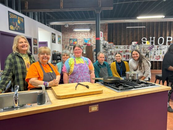 Group at a station in Thyme in Your Kitchen pose and smile for the camera together while the soup and chicken cook on the stove. 