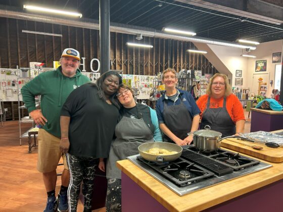Group at a station in Thyme in Your Kitchen pose and smile for the camera together while the soup and chicken cook on the stove. 