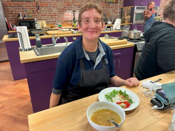 Photo of Caucasian attendee at Thyme in Your Kitchen wearing an apron and navy sport jacket sitting at the table with beautiful fresh made soup and chicken 