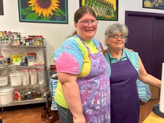 Photo of two attendees at Thyme in Your Kitchen. Both Caucasian females are wearing glasses and aprons but one is young with blonde-brown hair and the other is older with gray hair. 