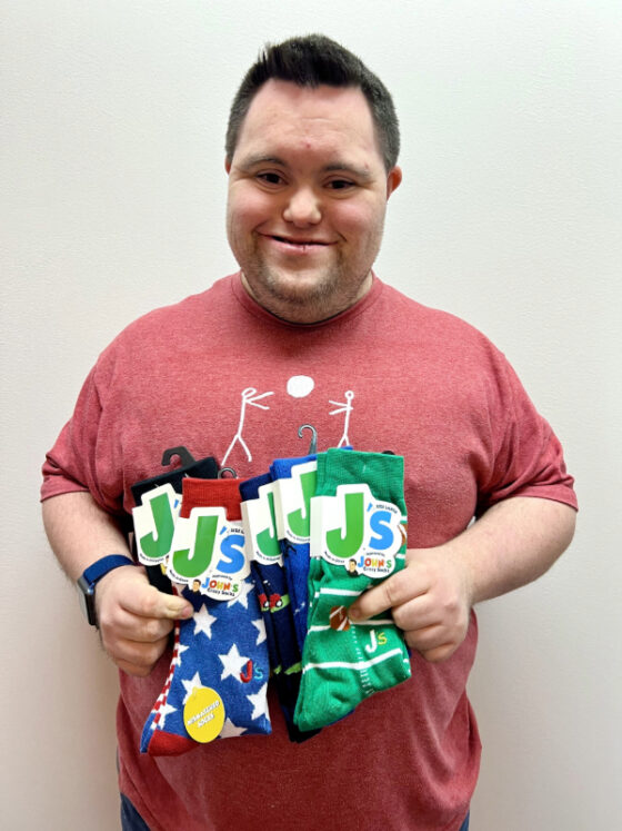 Photo of and provided by John Cronin, co-owner of John's Crazy Socks. He is wearing a red t-shirt, smiling, and holding five pair of bright, fun socks with logo and packaging. 