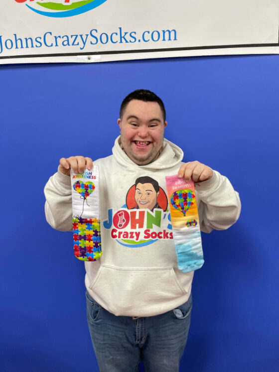 Photo of and provided by John, co-owner of John's Crazy Socks. He is wearing a white hoodie with his own logo and jeans. He is also smiling big and holding up two pairs of Autism advocacy awareness socks featuring colorful puzzle pieces. Behind him is a banner displaying the website JohnsCrazySocks.com