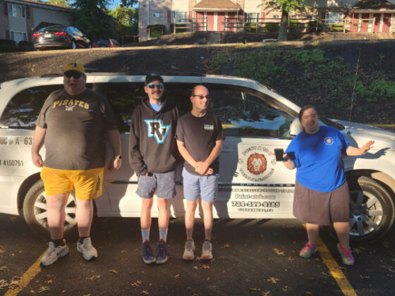 Photo of four people receiving transportation services standing in front of the white van on either side of the logo on the front door. They are all wearing shorts and t-shirts but do not match. The photo is light dappled as if it is a sunny day with a lot of nearby trees casting shadows. They are parked in a suburban community with leaves on the cement near their shoes. The image features the topic of transportation in rural areas. 