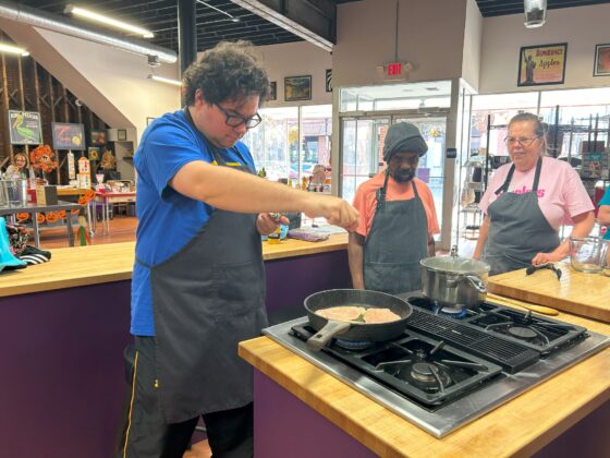 Attendees at Thyme in Your Kitchen. A Caucasian man in a blue t-shirt and apron is seasoning chicken in the skillet. A Caucasian women and a Black man are also wearing aprons watch with smiles and interest. 