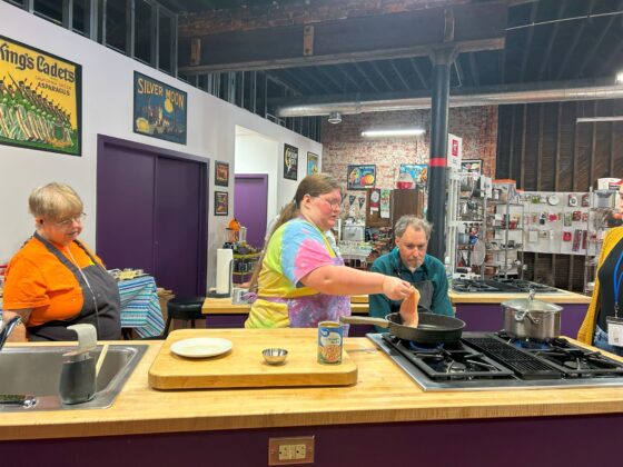 Attendees at Thyme in Your Kitchen, a Caucasian woman with long brown blonde hair pulled in a pony tail carefully places raw chicken in the skillet. A woman and man both Caucasian watch with interest. Everyone is wearing aprons. 