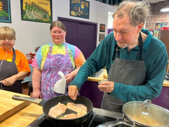 Attendees at Thyme in Your Kitchen, a Caucasian man carefully seasons chicken in the skillet. Two woman both Caucasian watch with interest. Everyone is wearing aprons. 