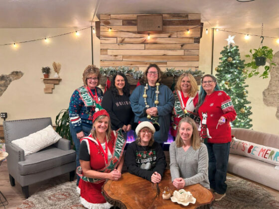 The photo features eight members of the Milestone HCQU West Team in a bright cozy room decorated for the holidays with lights, Christmas tree with a star, and a fireplace with garland. The women are wearing warm sweaters with a variety of festive patterns and are all facing the camera smiling. 