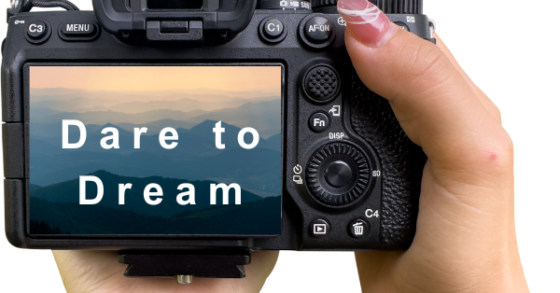 Image of a woman's hands with pink painted fingernails holding a camera where the screen display shows mountainous horizon with the text "Dare to Dream"