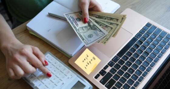 Photo of a caucasian woman's hands with red painted fingernails working at a desk with a computer, cash, and a calculator. A post it on the laptop shows the text "Budget Plan"