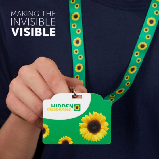 Image of someone in a navy blue t-shirt wearing the bold Sunflower green lanyard with a Sunflower card that reads "Hidden Disabilities" White text in teh corner says "Making the Invisible Visible." 