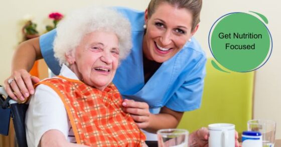 Image of an elderly woman in a wheelchair pushed up to the table with a glass of water and a white coffee mug. A younger woman in blue scrubs and white earrings is assisting her with a checkered bib to protect her clothes from potential food or drink spills. Both women are Caucasian and have big smiles toward the camera. A green circle text box reads "Get Nutrition Focused." 
