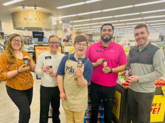Photo of Max wearing a personalized apron and standing with employees all holding up Max Mix bottles. 