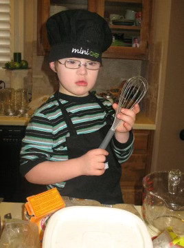 Photo of young Max wearing a chef hat, holding a whisk and working in the kitchen. 