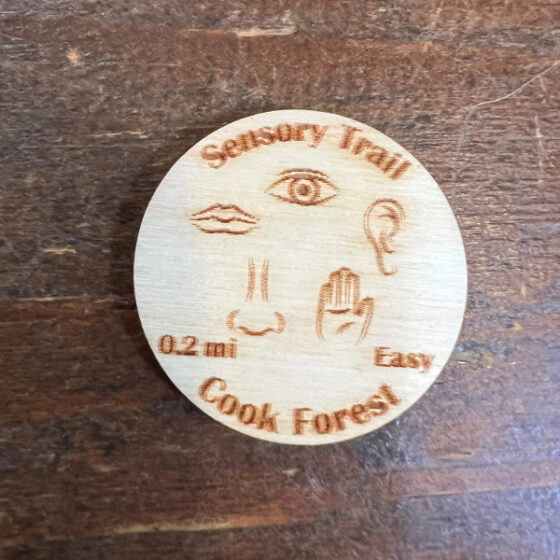 Souvenir wooden coin for the Cook Forest 0.2 mi easy Sensory Trail. Besides the title Icons are shown clockwise for eye, ear, hand, nose, and mouth.
